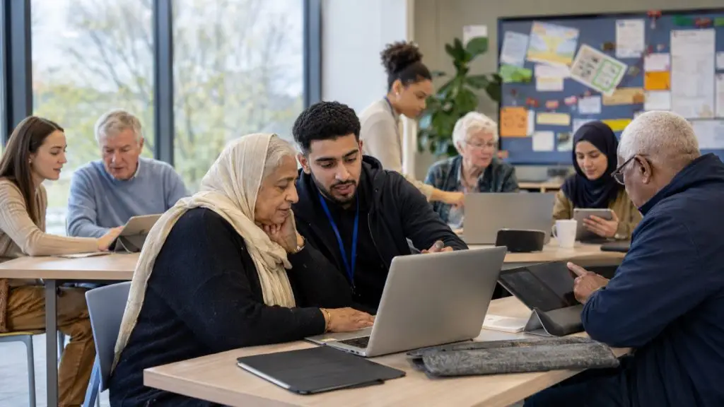 A young digital champion helping an older woman use a laptop at a busy, diverse community digital inclusion session